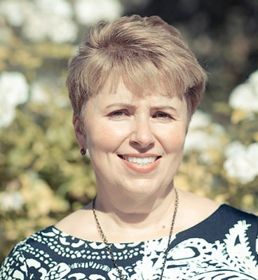 This is a portrait of an older woman with short hair, smiling at the camera. She wears glasses and has a necklace around her neck. The background is out of focus but appears to be an outdoor setting with greenery.