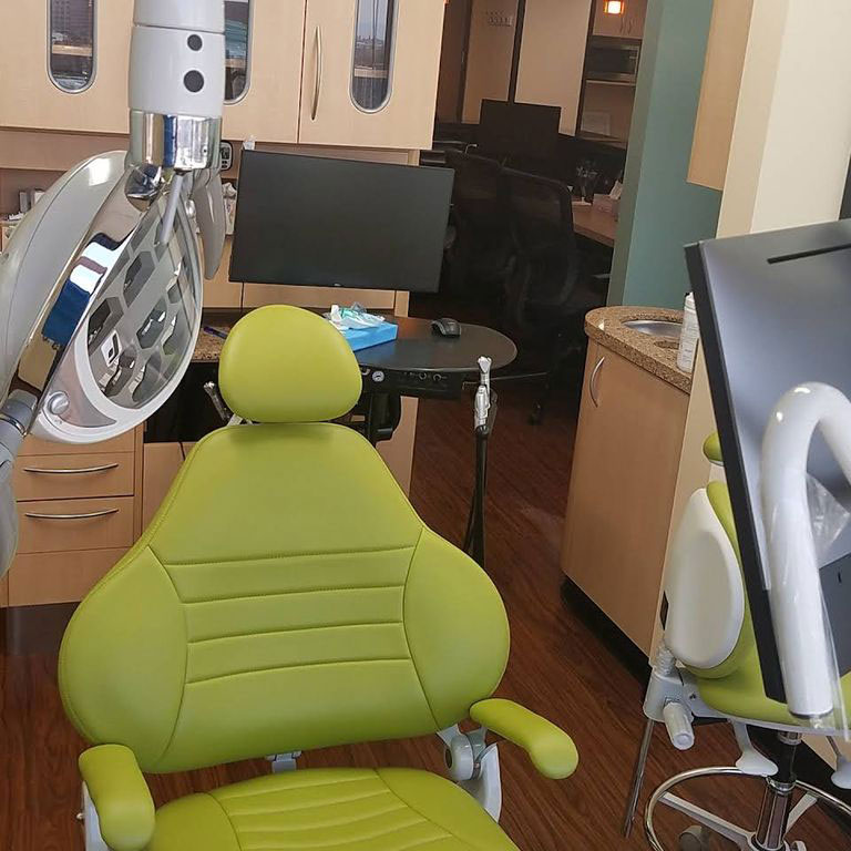 The image shows a dental office with a modern chair, a computer monitor, and a desk area in the background.