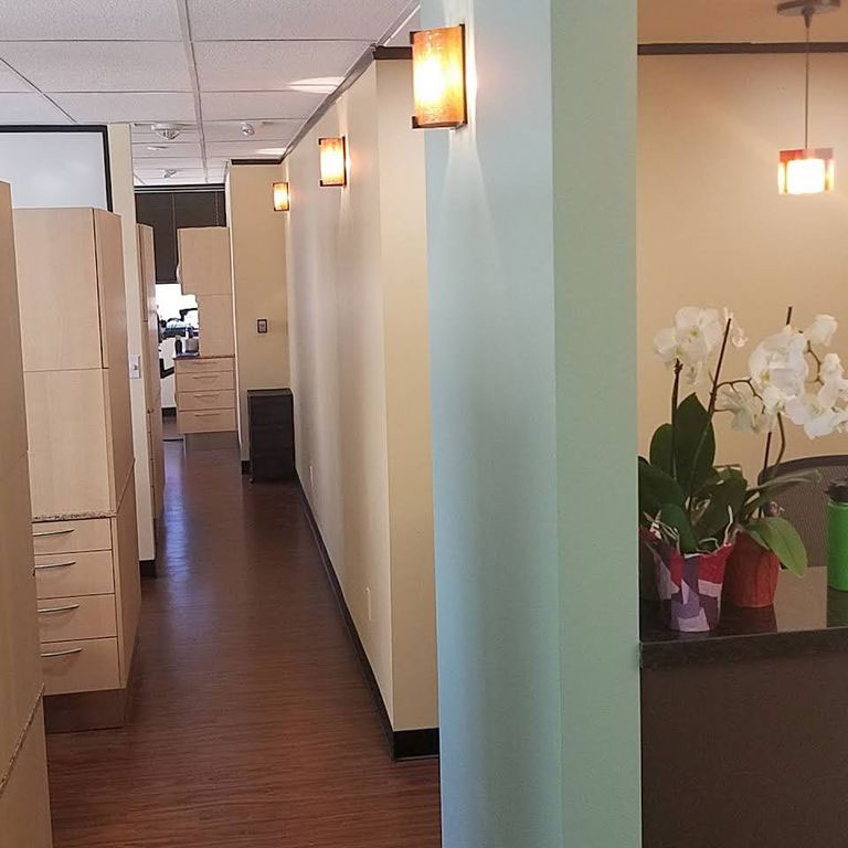 The image shows an interior space that appears to be an office or waiting area with a long hallway leading to a reception desk, featuring light-colored walls, greenish flooring, and modern lighting fixtures.