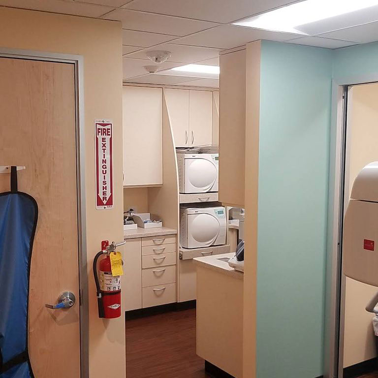 The image depicts an interior view of a room with a kitchenette area featuring cabinets and appliances, adjacent to a hallway leading to a restroom with a sink and a mirror.