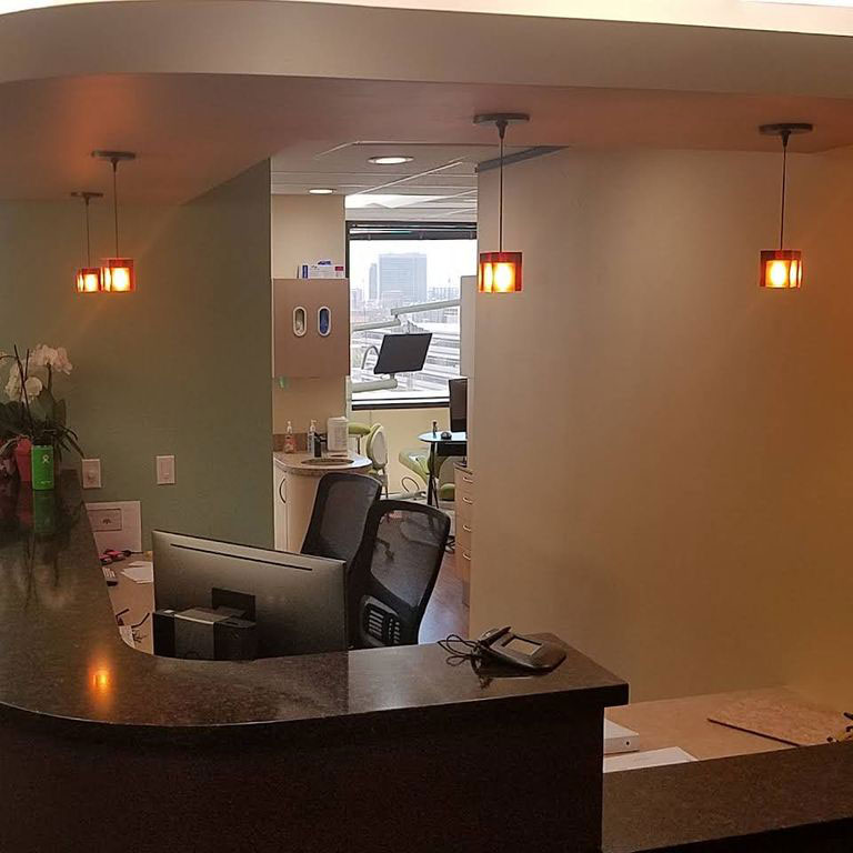 The image shows an interior view of a modern office space with a reception area featuring a counter, chairs, and lighting fixtures, set against a backdrop of a wall-mounted TV and a window with a cityscape visible beyond.