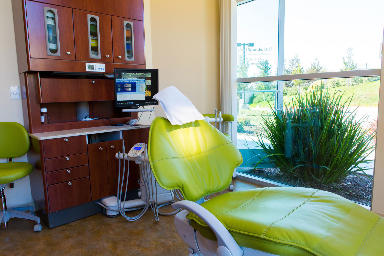 The image shows a dental office with a green dental chair and a desk with a computer monitor, situated next to a window.