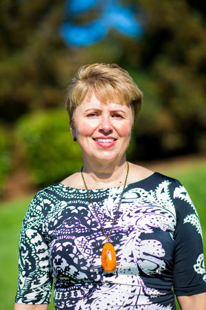 A woman stands outdoors, smiling at the camera, wearing a patterned top with a necklace and a lanyard.