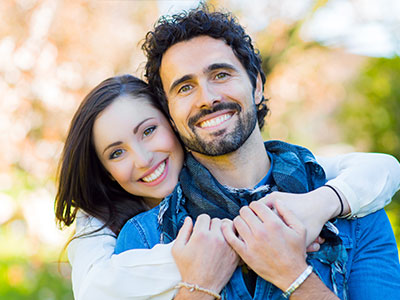 Man and woman embracing  man has beard, woman has long hair, both are smiling, wearing casual clothing, outdoors, daylight.