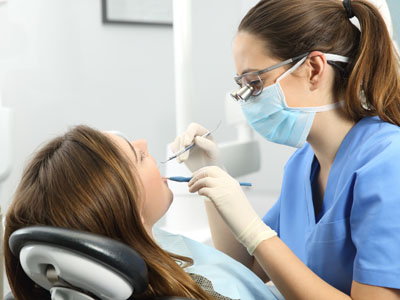 A dental hygienist is assisting a woman with her dental care, both wearing face masks and sitting in a dental office chair.