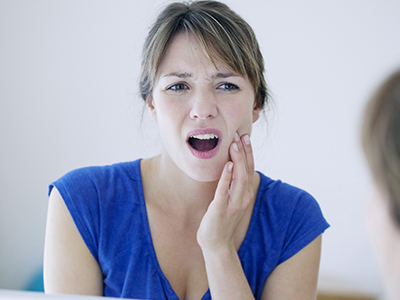 An adult female with a concerned expression and her mouth open, looking into a mirror while holding her hand on her face.