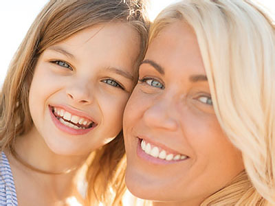 The image shows a woman with blonde hair and a young girl with brown hair smiling at the camera. They appear to be outdoors, possibly on a beach, given the sunlight and bright colors.