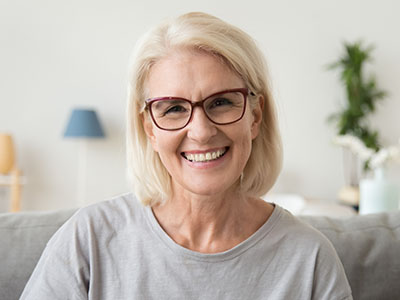 The image shows a woman with short blonde hair smiling at the camera. She has glasses on her face, wears a light-colored top, and is seated indoors with a blurred background that includes furniture and what appears to be a lamp.