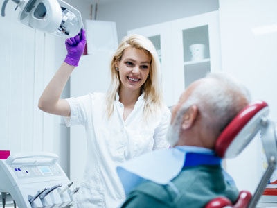 A woman with blonde hair is standing next to an older man who is seated in a dental chair, being attended to by a dental professional.