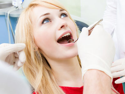 The image shows a woman sitting in a dental chair with her mouth open, receiving dental care from a professional wearing protective gloves and a face mask.