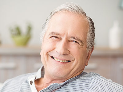 The image shows an older man with white hair, smiling and looking directly at the camera. He appears to be seated indoors, possibly in a living room setting, and has a relaxed posture.