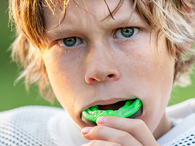 The image shows a young person with blonde hair holding a green object in their mouth while looking directly at the camera.