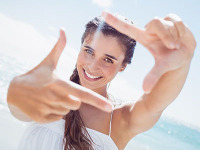 A woman with long hair and a white top takes a selfie with her hand held up to the camera while standing on a beach.